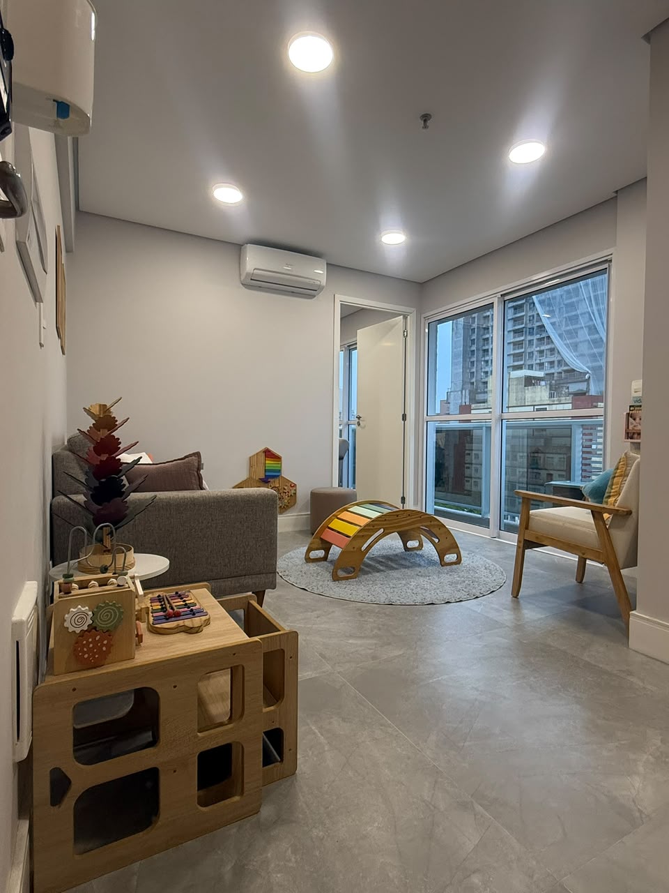 Sala de espera lúdica com brinquedos de madeira, arco montessoriano colorido e janelas amplas